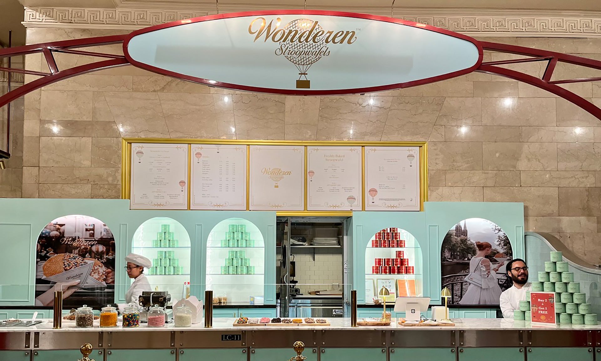 Interior view of the Wonderen Stroopwafels store with an elegant marble backdrop and a pastel blue counter. Large menu boards hang above showcasing the freshly baked stroopwafel options. Two employees are present, one wearing a chef's hat busy with food preparation and the other smiling at the camera. Stacks of stroopwafel boxes in varying colors are neatly arranged on the counter, with a display of loose wafels and toppings in front. The store emanates a welcoming and luxurious atmosphere.
