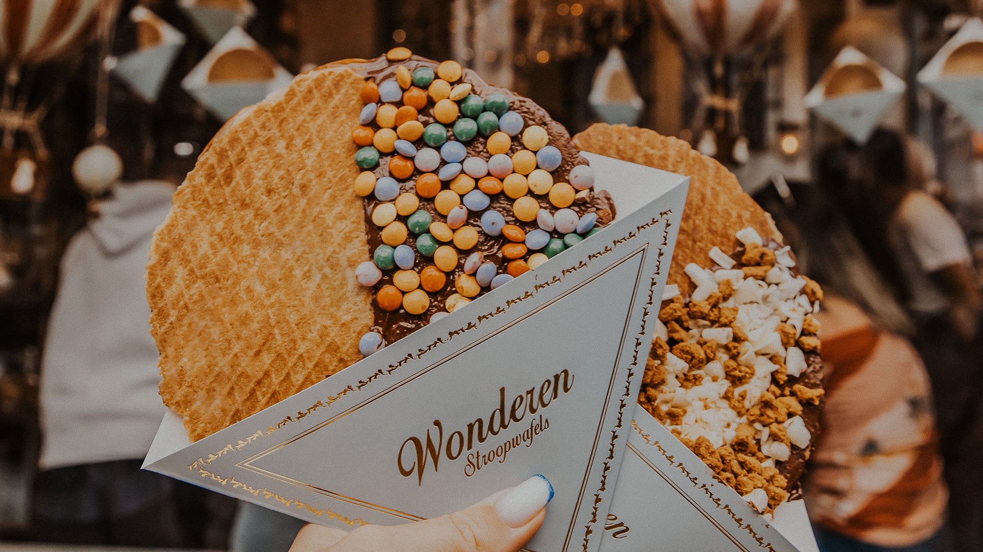 A close-up of two hands holding a Wonderen Stroopwafel, each wafel generously topped with colorful treats. The left one is covered with a vibrant mix of multi-colored coated chocolates, while the right one is sprinkled with white mini marshmallows and crumbled biscuit. They are presented in branded Wonderen Stroopwafels sleeves with elegant gold detailing. In the blurred background, the warm ambiance of the store with its lighting and other customers is visible.
